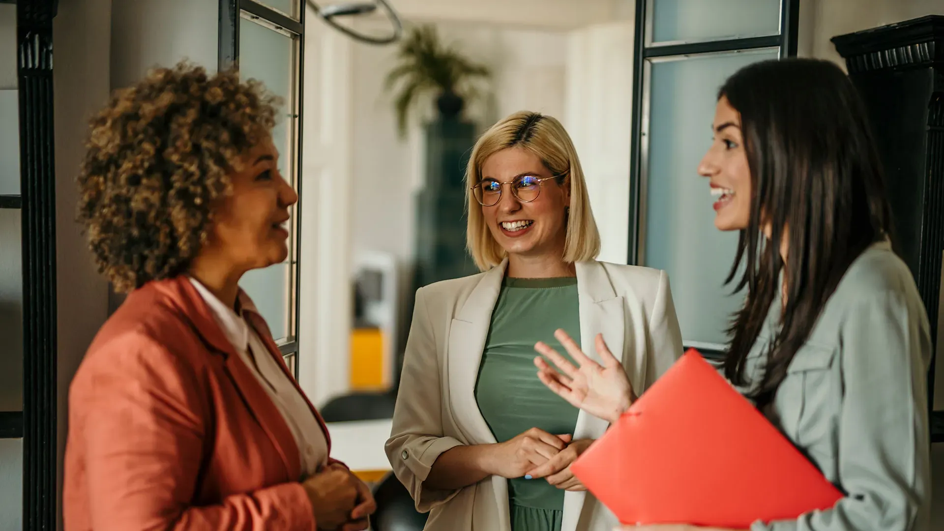 Three women engaged in conversation in an office hallway, with a professional atmosphere and neutral decor
